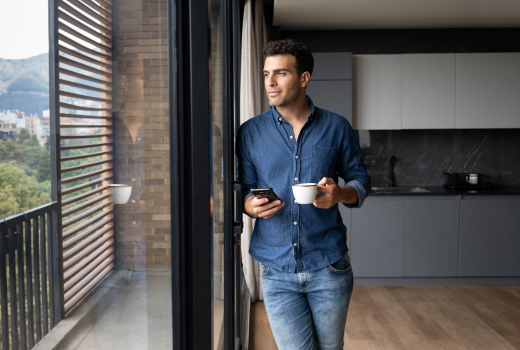 PIA Eine Person, steht in einer modernen Küche und hält eine Tasse Kaffee in der einen Hand und ein Smartphone in der anderen. Im Hintergrund sind große Fenster mit Blick auf eine Landschaft mit Bäumen und Bergen zu sehen.