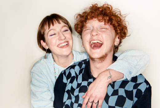 PIA Zwei Personen, posieren vor einem neutralen Hintergrund. Eine Person hat den Arm um die Schulter der anderen gelegt.