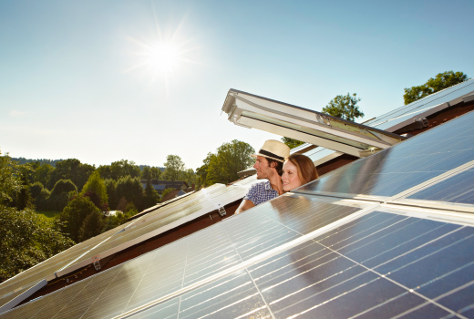 PIA Zwei Personen, schauen aus einem Dachfenster heraus auf ein Dach mit Solarpaneelen. Im Hintergrund sind Bäume und ein klarer Himmel zu sehen.