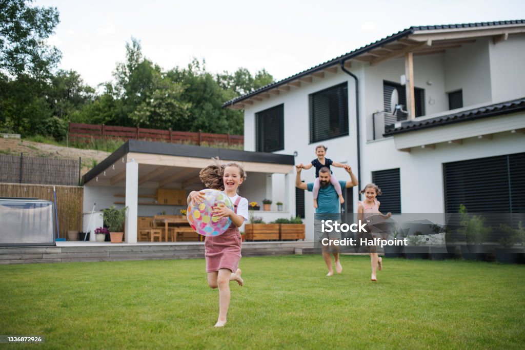 Familie spielt fröhlich im Garten vor einem modernen Haus – ein Kind läuft mit einem Ball, die anderen folgen lachend.
